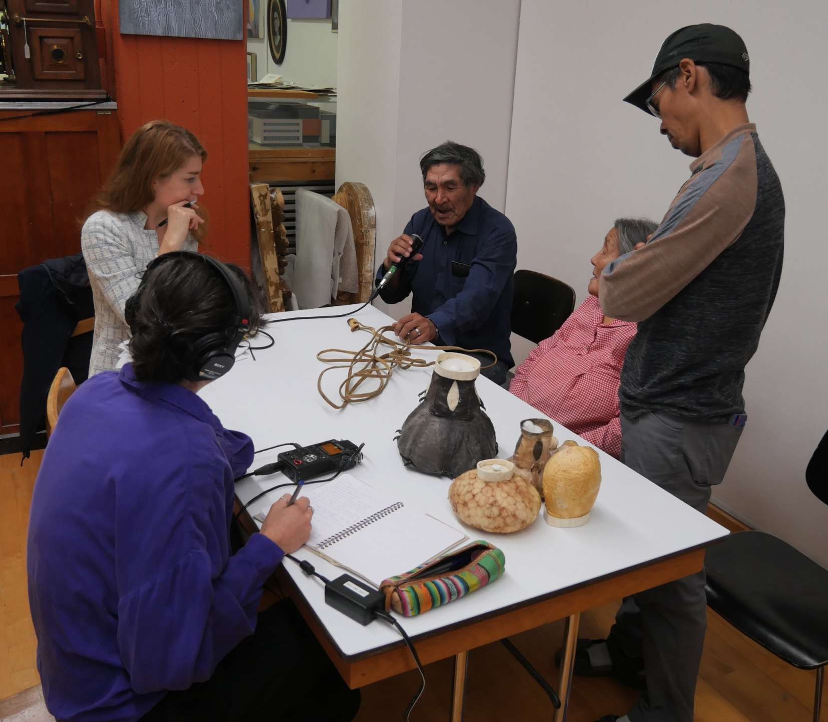 Cinq personnes sont réunis autour d'une table, sur laquelle sont posés plusieurs objets inuit. Un ainé inuk a un micro dans une main et tient une corde de tête de harpon dans l'autre main. Une autre personne a un casque sur les oreilles relié à une enregistreuse.