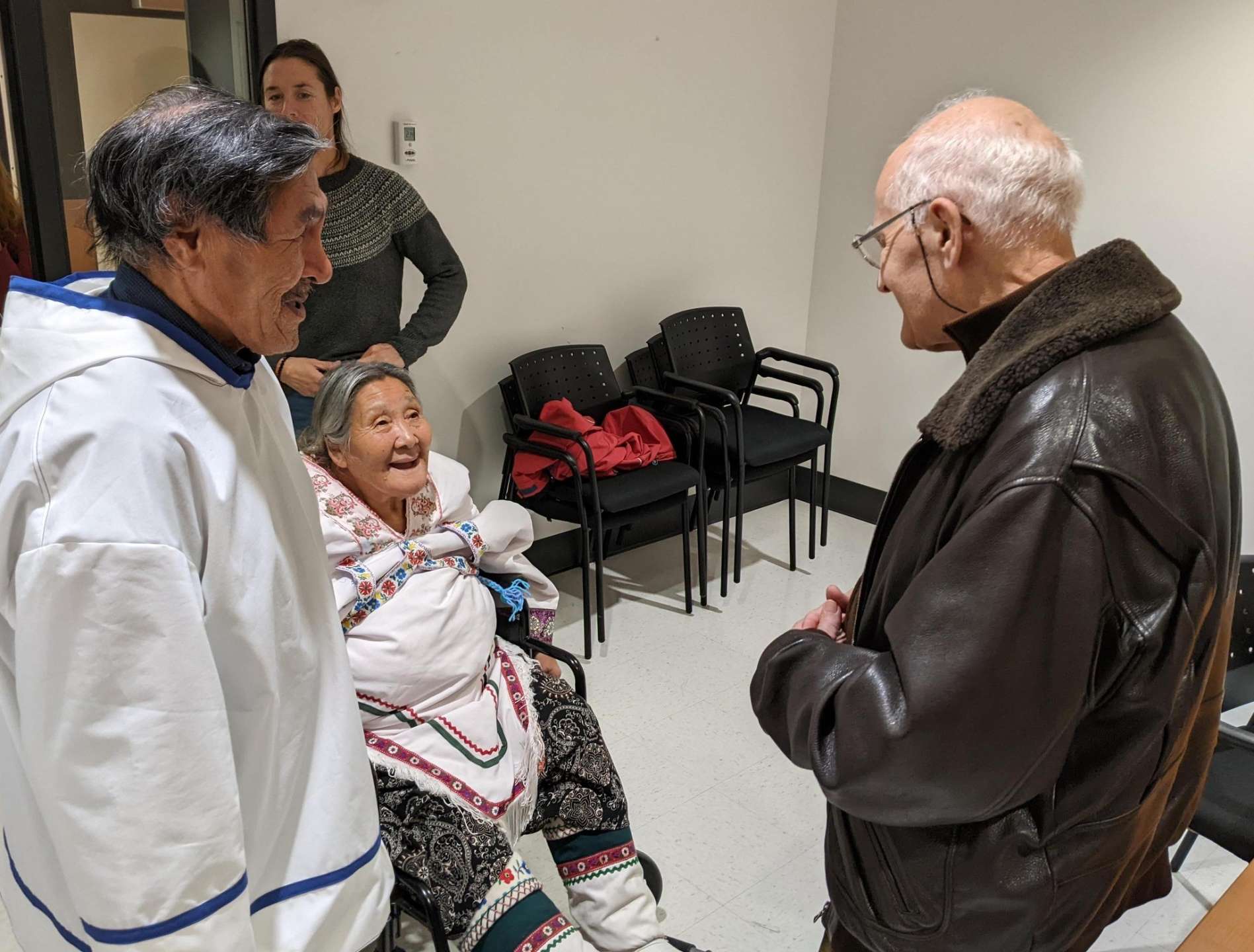 Bernard Saladin d'Anglure se tient debout face à Deborah Qaunaq (ainée inuk, assise) et Natalino Piugattuk (ainé inuk, debout). Les deux ainés portent un amauti blanc avec de fines décorations de couleurs. Tous sourient.