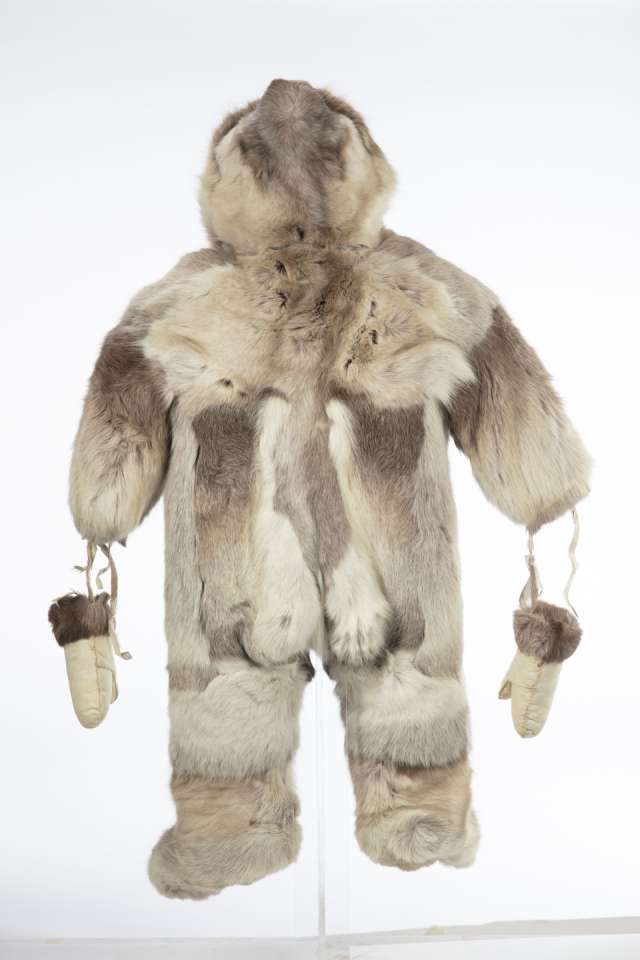 Combinaison en fourrure de caribou, vue de derri&egrave;re, avec le capuchon relev&eacute;. Des mitaines de cuir retourn&eacute;, orn&eacute;es de fourrure brune aux poignets, sont suspendues aux manches par des lani&egrave;res. La fourrure pr&eacute;sente diff&eacute;rentes nuances de brun clair et fonc&eacute;.