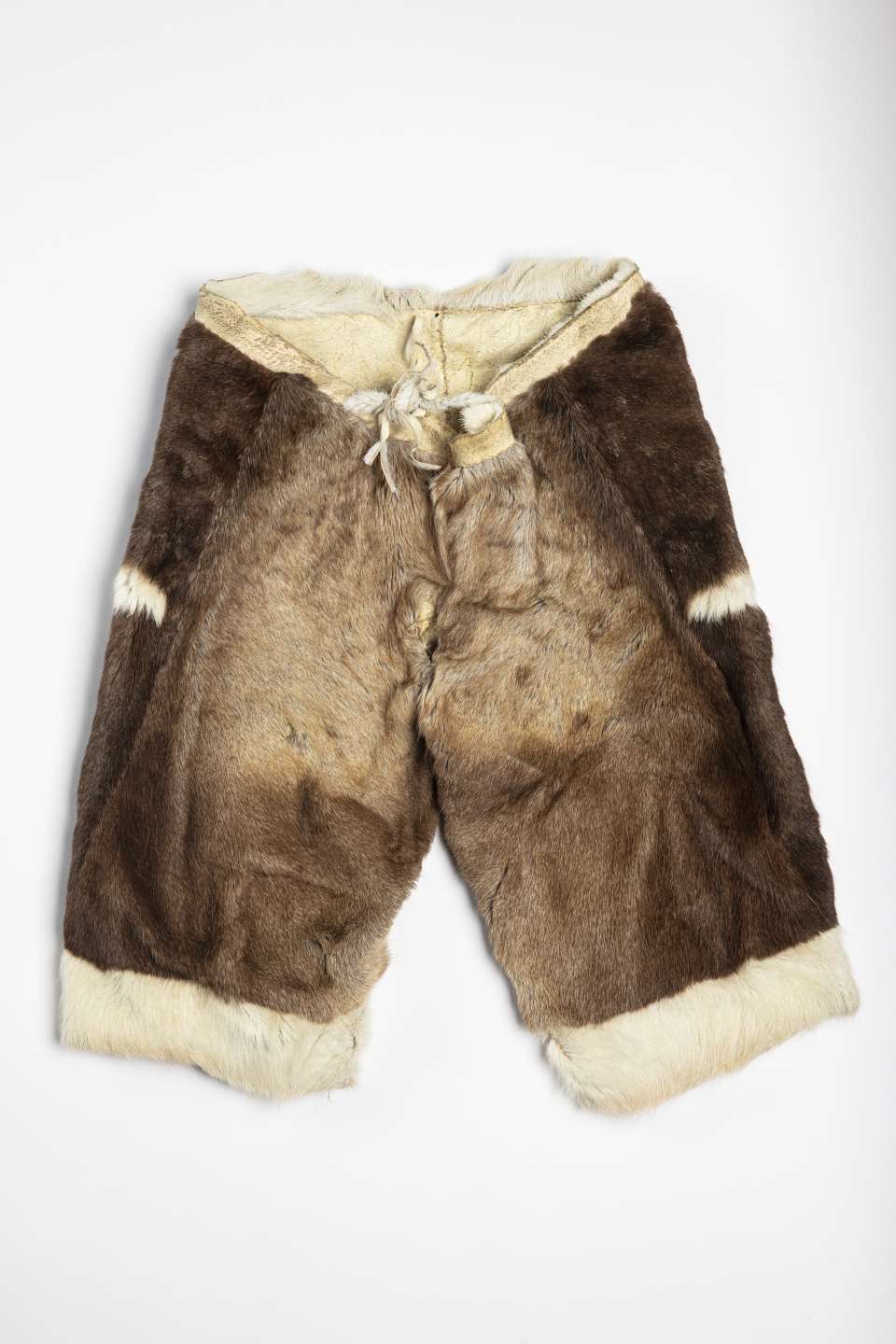 Pantalon en fourrure brune, vu de face, avec deux courtes bandes blanches sur les c&ocirc;t&eacute;s et une bande de fourrure blanche au bas de chaque jambe. La taille en cuir retourn&eacute; est ferm&eacute;e par un cordon nou&eacute; &agrave; l&rsquo;arri&egrave;re, mais visible &agrave; l&rsquo;avant.