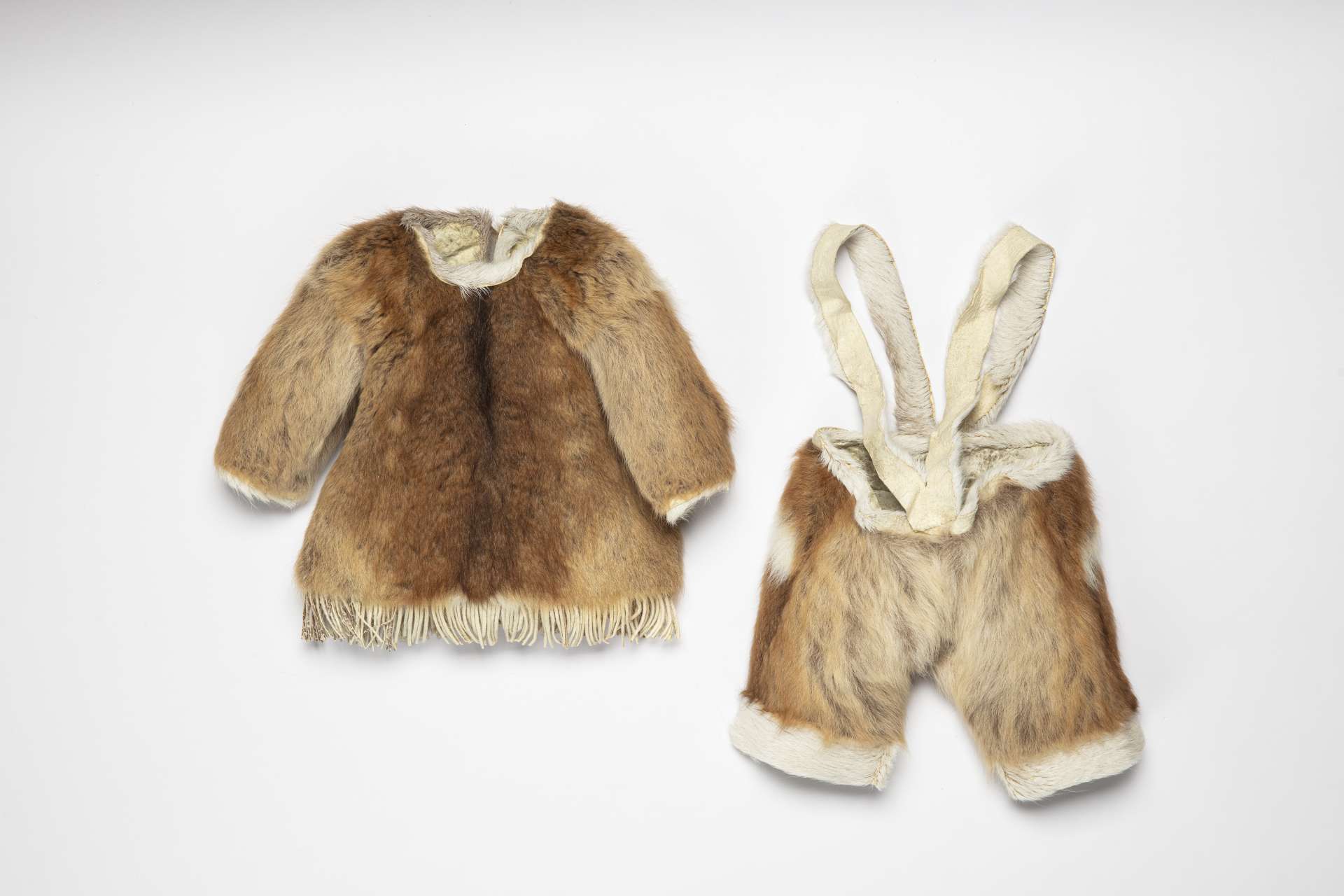 Petit ensemble en fourrure, vu de devant, compos&eacute; d&rsquo;un manteau et d&rsquo;un pantalon &agrave; bretelles. Le manteau est dot&eacute; de franges en bas. Une bande de fourrure blanche borde la taille et le bas de chaque jambe du pantalon, qui est dot&eacute; de larges bretelles en peau.