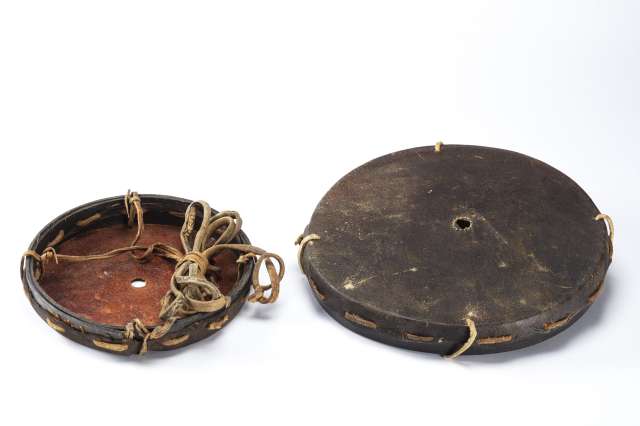 Deux objets circulaires &agrave; bords &eacute;pais, en cuir brun fonc&eacute;, tendu sur un cerclage. Tous les deux pr&eacute;sentant un trou &eacute;troit au centre. L&rsquo;un est renvers&eacute; montrant l&rsquo;int&eacute;rieur rouge&acirc;tre et son cordage en cuir brun attach&eacute; en quatre points au cerclage.