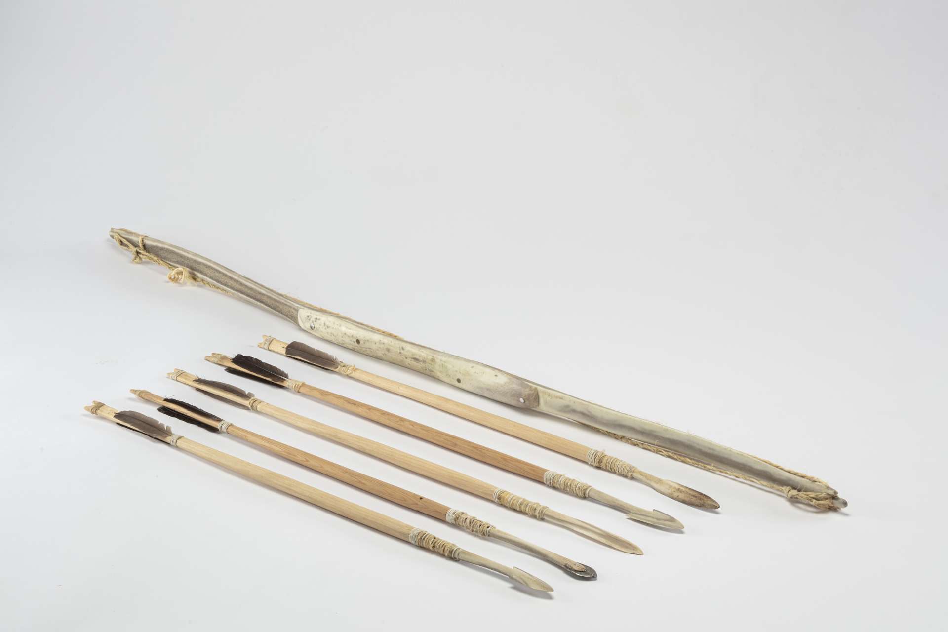 Un arc en mati&egrave;re osseuse blanche accompagn&eacute; de cinq fl&egrave;ches en bois avec des plumes et des pointes de diff&eacute;rentes formes, en m&eacute;tal et en os.