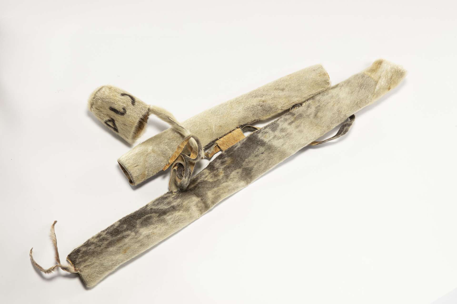 Deux &eacute;tuis cylindriques dispos&eacute;s l&rsquo;un &agrave; c&ocirc;t&eacute; de l&rsquo;autre. Le premier est en peau de phoque sombre avec des taches plus claires en forme d&rsquo;anneaux. Le second est plus court et en fourrure beige p&acirc;le, reli&eacute; &agrave; son capuchon par une lani&egrave;re enroul&eacute;e, sur lequel est &eacute;crit, en fourrure brune, &laquo; ugatu &raquo; en inuktitut syllabaire.