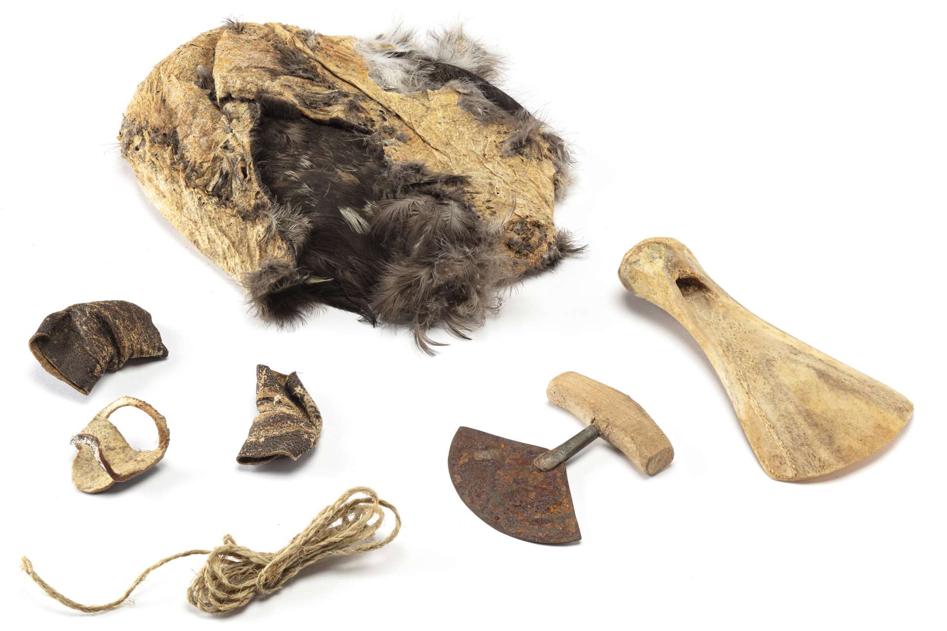 Vue de plusieurs outils de couture dispos&eacute;s en cercle, comprenant une trousse en peau de guillemot retourn&eacute;e, un couteau en demi-lune avec une lame rouill&eacute;e, un grattoir en os de forme allong&eacute;e et incurv&eacute;e, trois petites pi&egrave;ces en cuir et une corde tress&eacute;e de couleur beige.