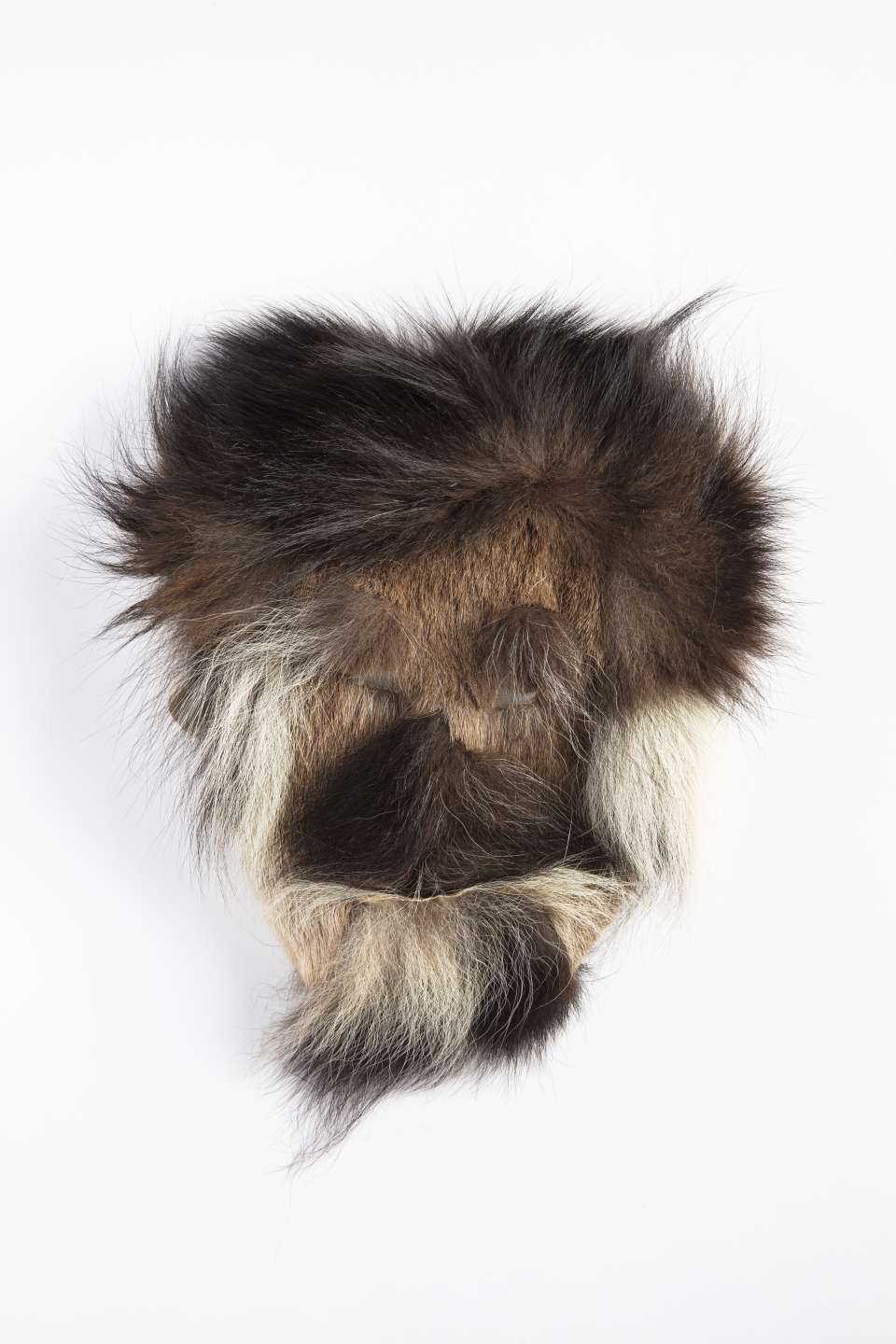Masque en fourrure brune, vu de face, orn&eacute; de fourrure sombre, avec des touffes de fourrure noire et brune de part et d&rsquo;autre, et des zones plus claires sous le menton et autour des joues. La fourrure forme des lignes distinctes cr&eacute;ant une impression de cheveux, de barbe et de moustaches.