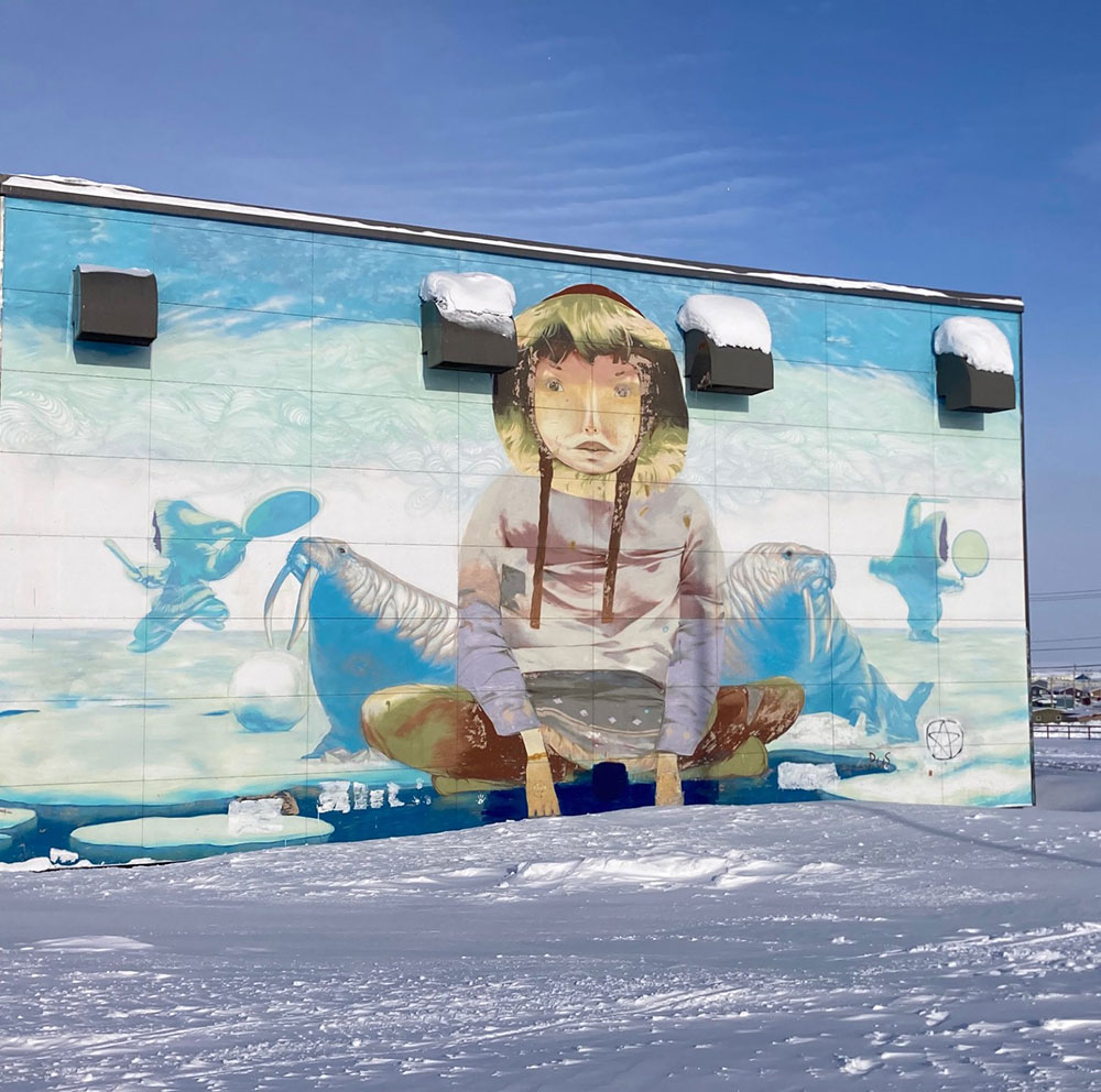 Igloolik en hiver : il y a de la neige au sol, une murale sur un bâtiment représentant un enfant assis, des morses et des danseurs.