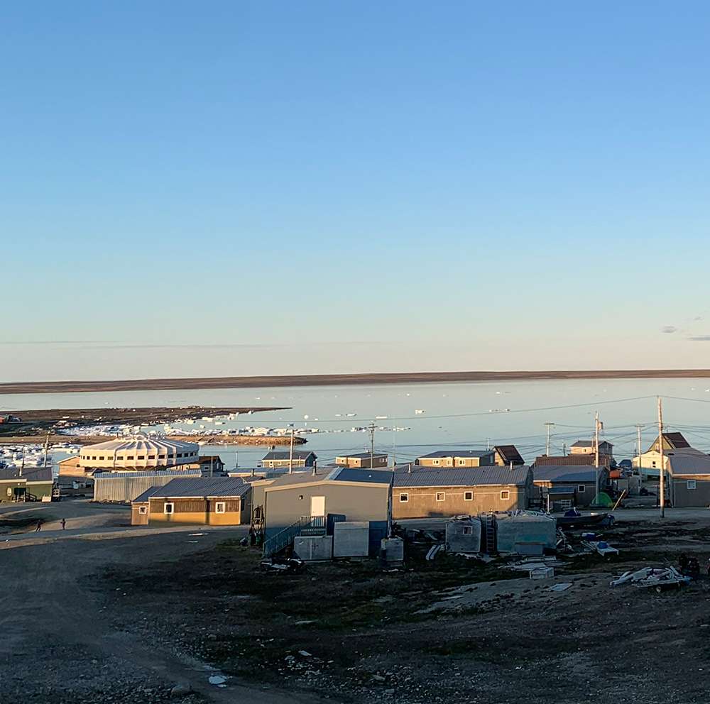 Vue d'Igloolik en été: pas de neige au sol, plusieurs maisons toutes situées au bord de l'eau, un bâtiment en forme de champignon au milieu de celles-ci, et quelques morceaux de glace éparses au bord du rivage.